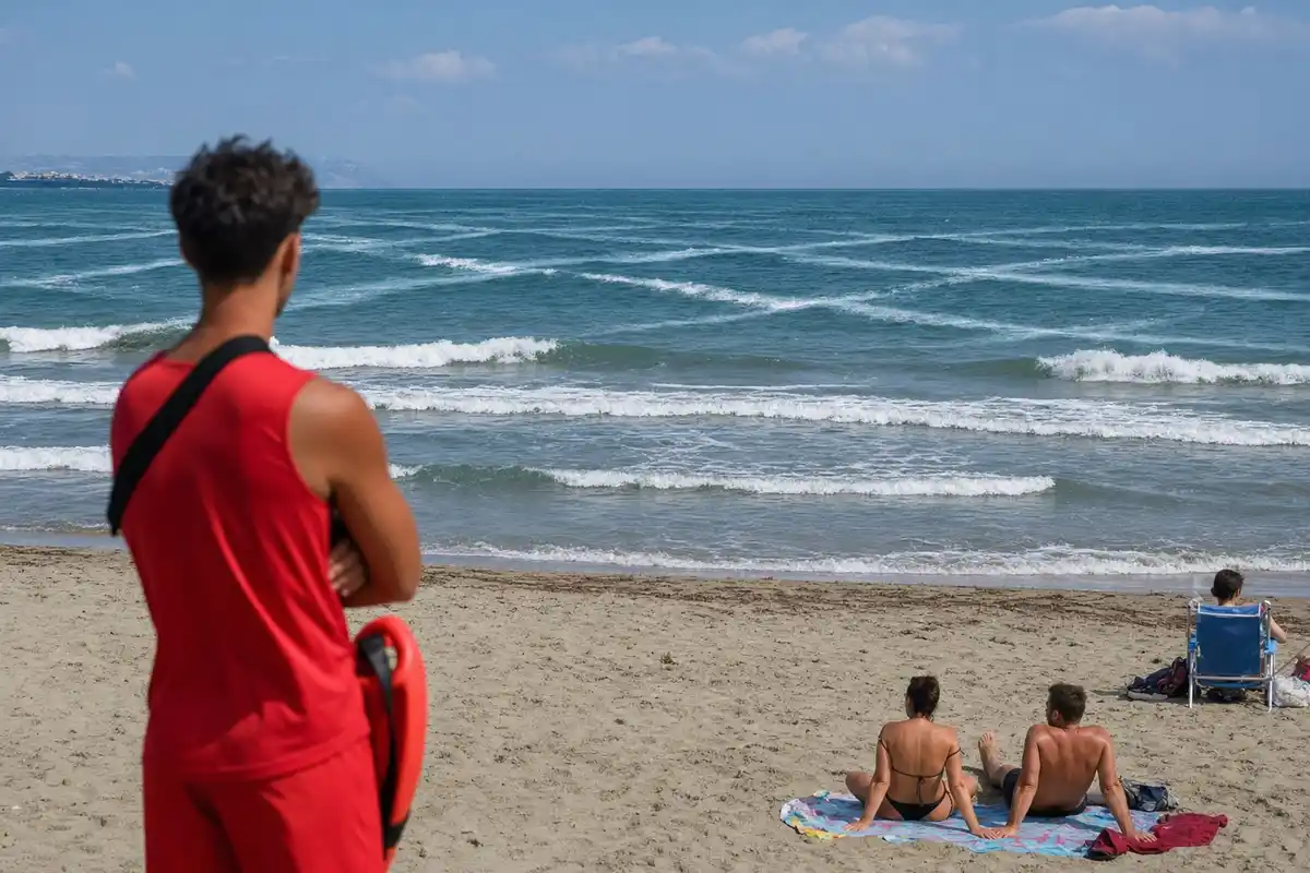 Bagnino in rosso osserva il mare con onde incrociate; alcuni bagnanti seduti sulla spiaggia