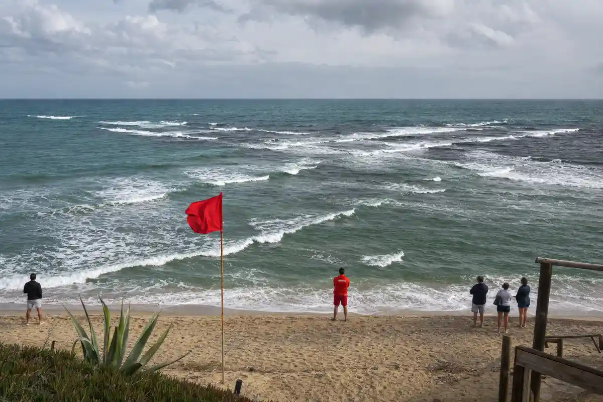 Bandiera rossa sulla spiaggia e mare mosso con onde incrociate; alcune persone osservano la riva sotto un cielo nuvoloso