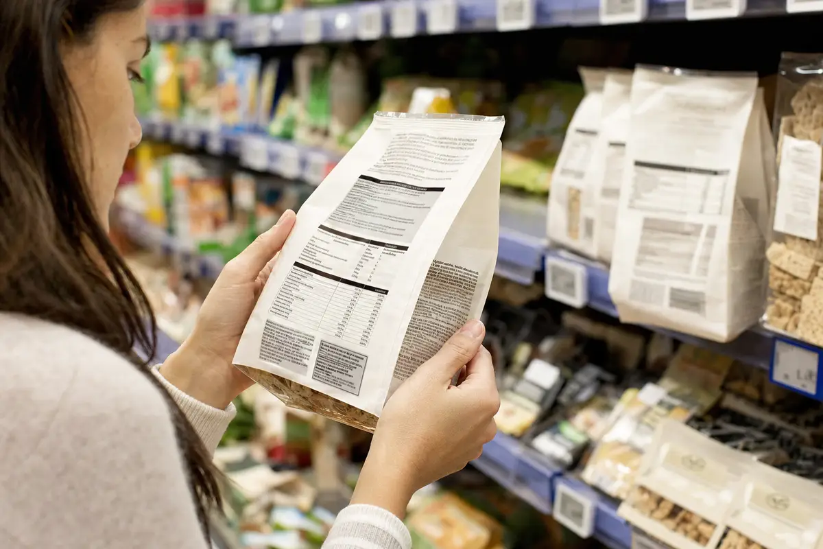 Cliente al supermercato legge l’etichetta nutrizionale sul retro di una confezione, con scaffali sullo sfondo