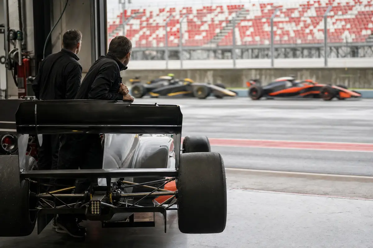 Meccanici in tuta nera osservano dal box una monoposto ferma mentre due auto sfrecciano sfocate in pit lane