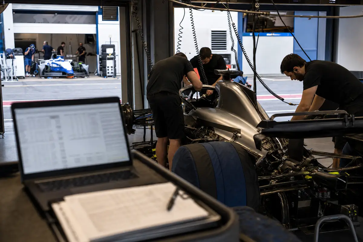 Meccanici al lavoro su una monoposto nel box, con laptop e documenti sfocati in primo piano