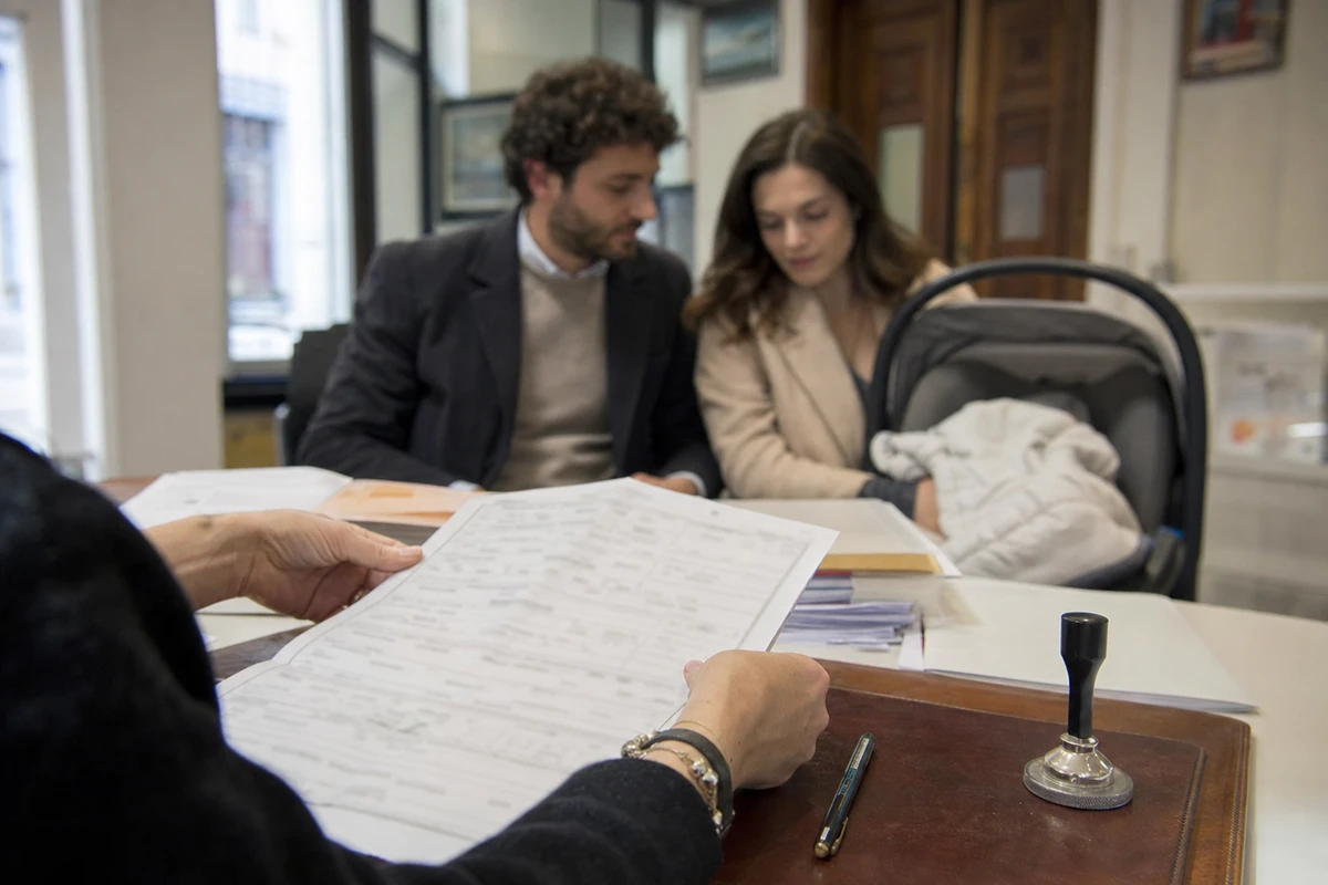 Impiegato con moduli e timbro su una scrivania, coppia con neonato in carrozzina sullo sfondo