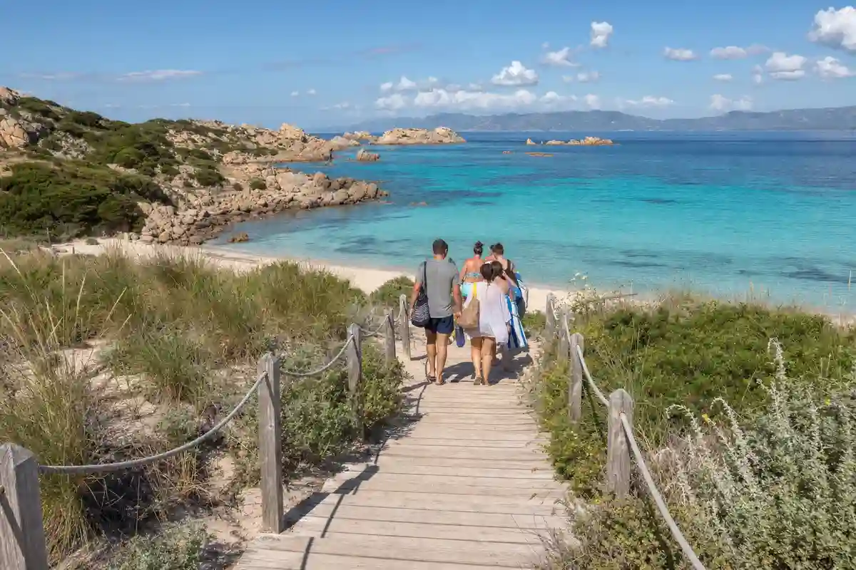 Passerella in legno tra le dune verso una spiaggia di sabbia bianca e mare turchese, con bagnanti di spalle