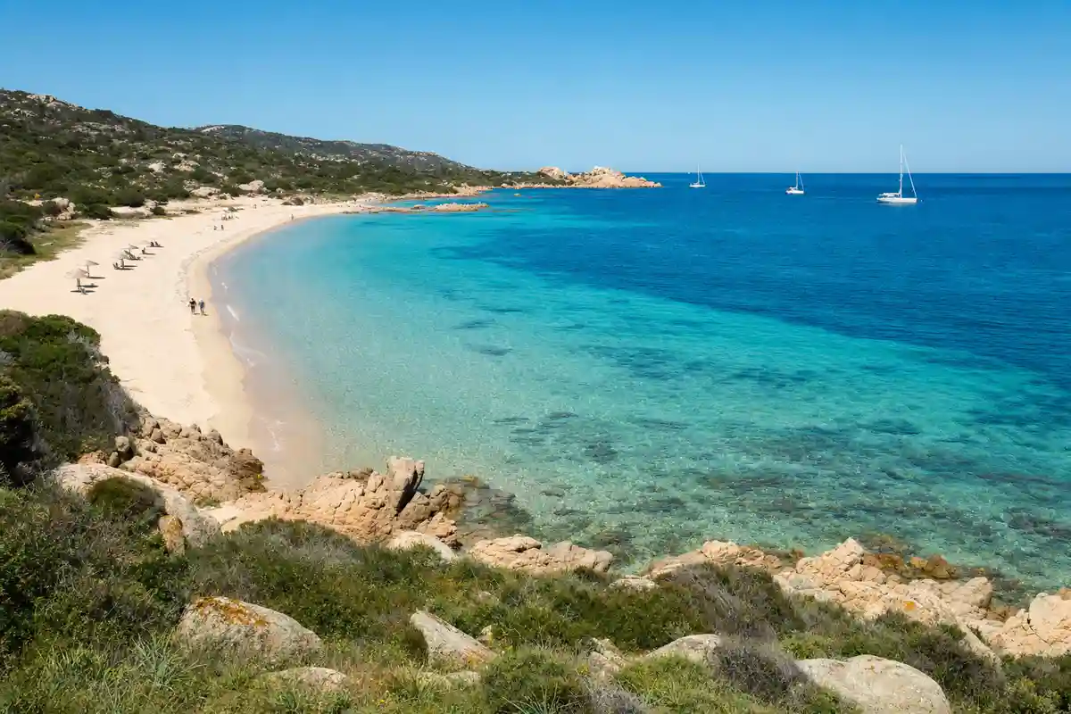Baia con spiaggia di sabbia chiara, acqua turchese trasparente, rocce e vegetazione mediterranea, barche a vela al largo