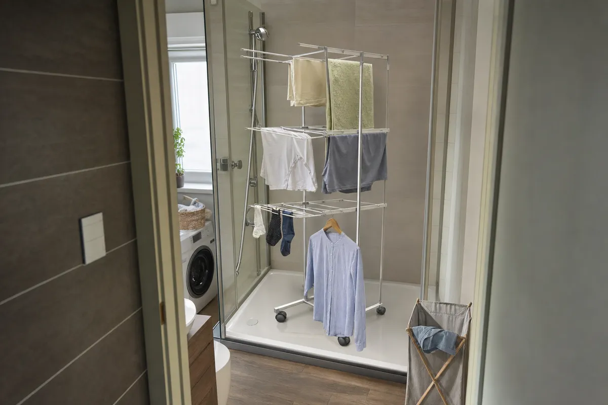 Stendino verticale a torre nella doccia con asciugamani, magliette e calze, bagno piccolo con lavatrice sullo sfondo