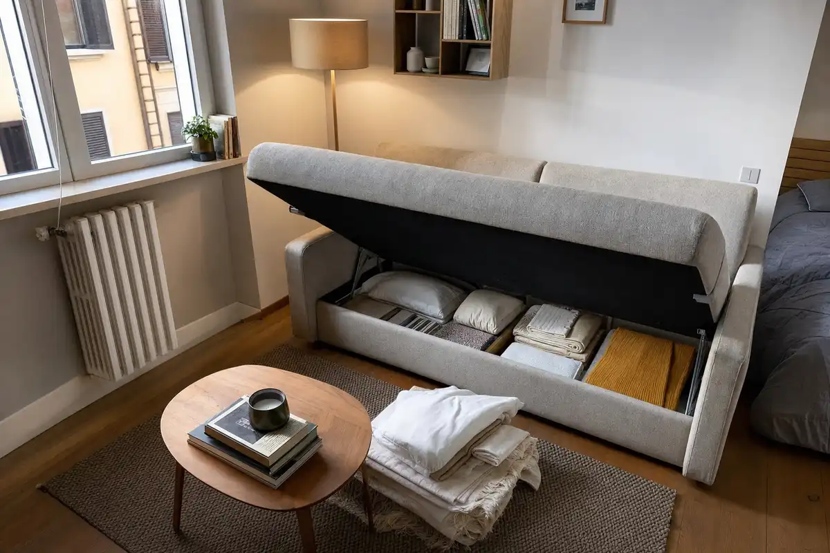 Divano letto grigio con seduta sollevata e vano contenitore pieno di coperte e cuscini in un monolocale