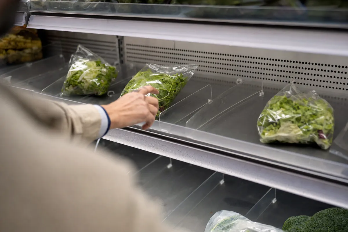 Scaffale frigo quasi vuoto con poche buste trasparenti di insalata pronta e una mano che ne prende una