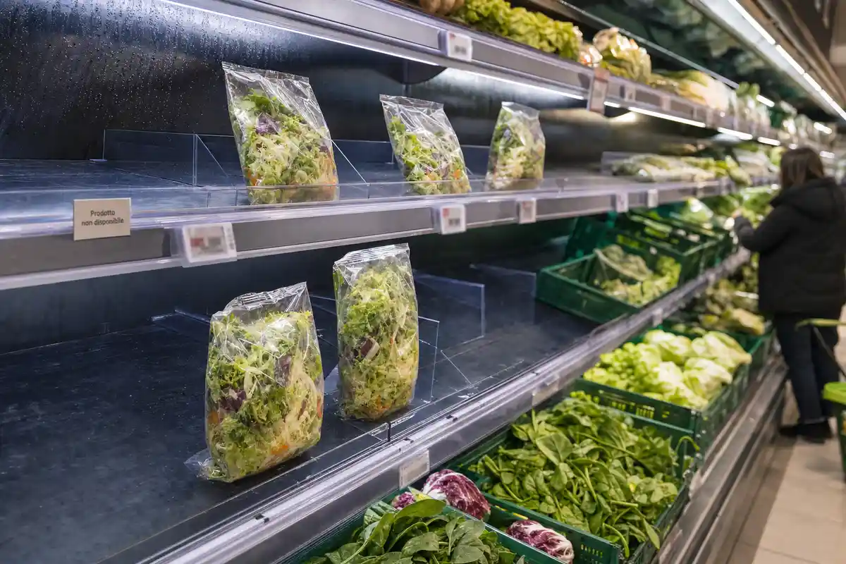 Scaffale frigo quasi vuoto con poche buste di insalata e cartello “Prodotto non disponibile” in supermercato