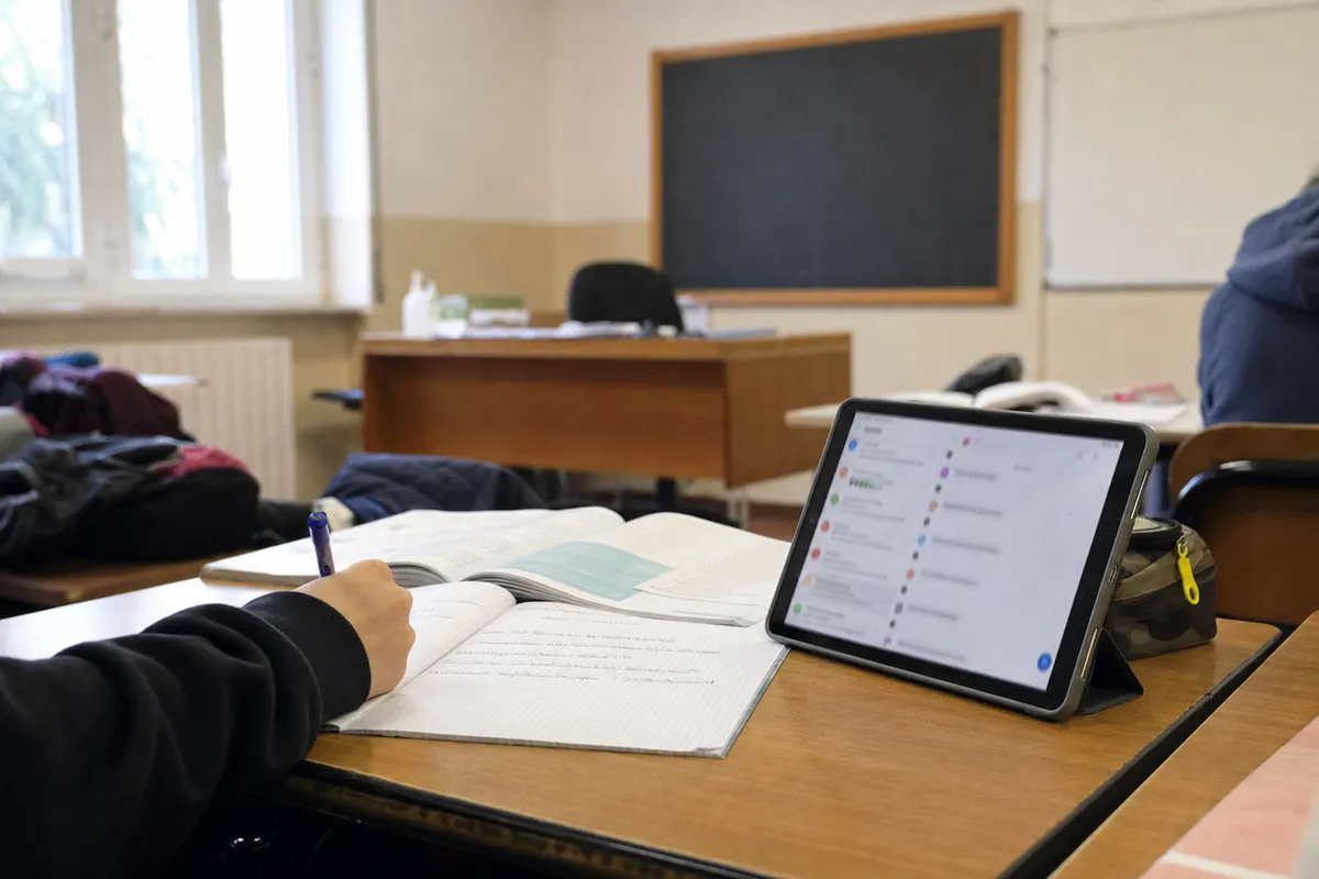 Banco in aula con quaderno aperto, mano che scrive e tablet con notifiche del registro elettronico sfocate