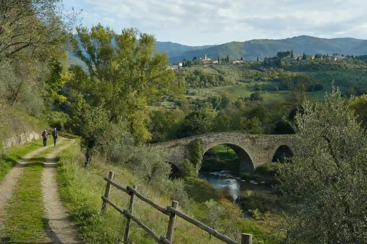 Sentiero di campagna con due escursionisti verso un ponte in pietra sul torrente, tra colline verdi del Mugello