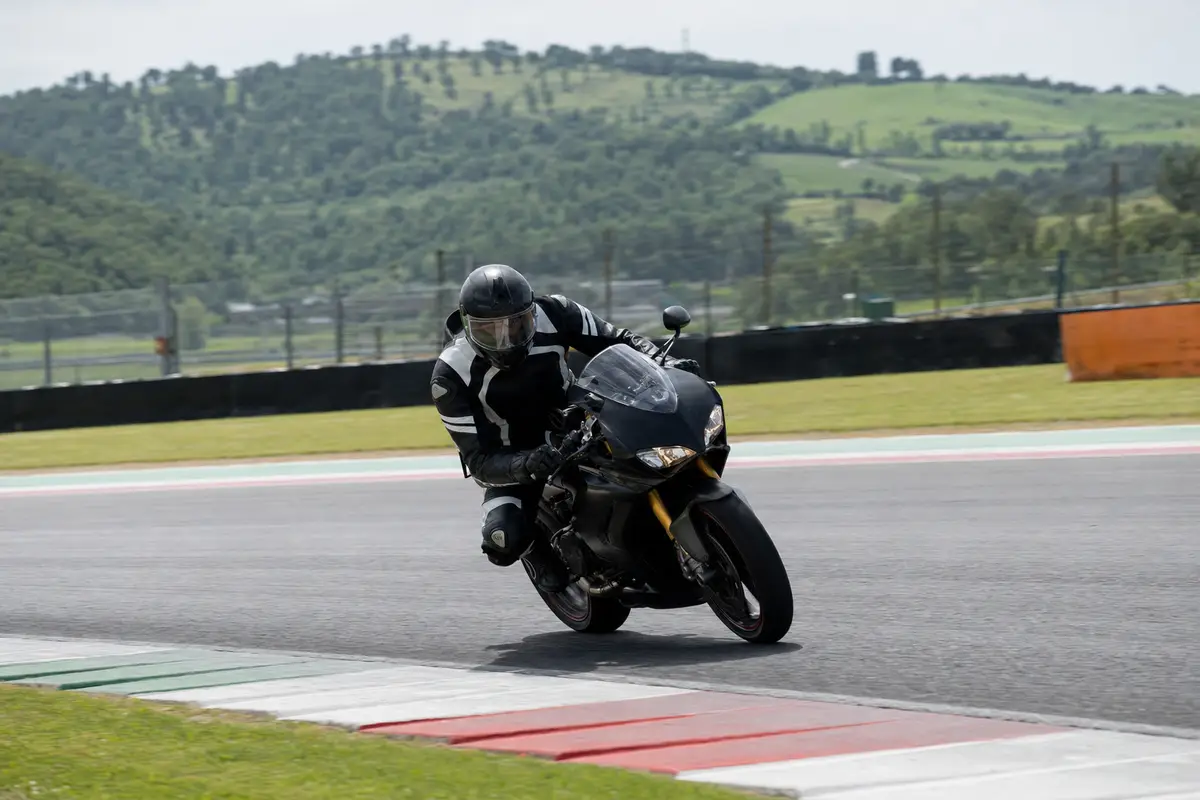 Motociclista in tuta protettiva piegato in curva su moto sportiva nera in pista, con colline verdi sullo sfondo