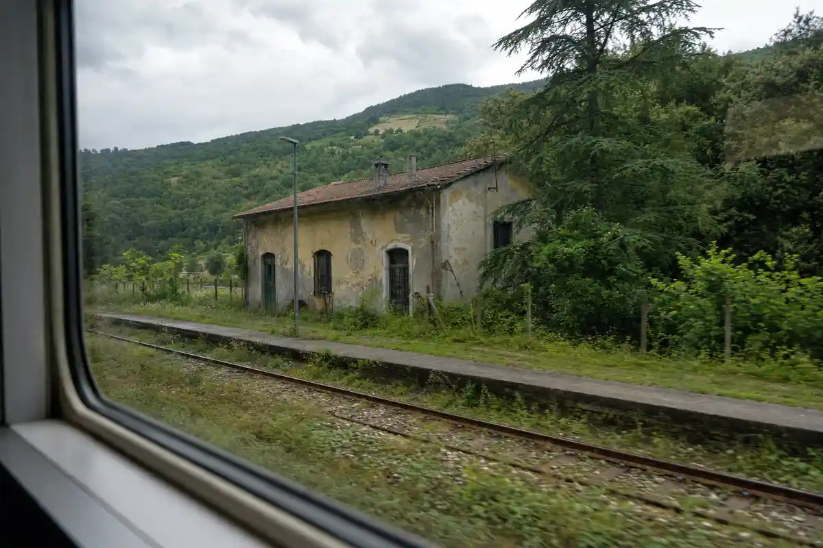 Vista dal finestrino di un treno: piccola stazione rurale con edificio scrostato tra verde e colline