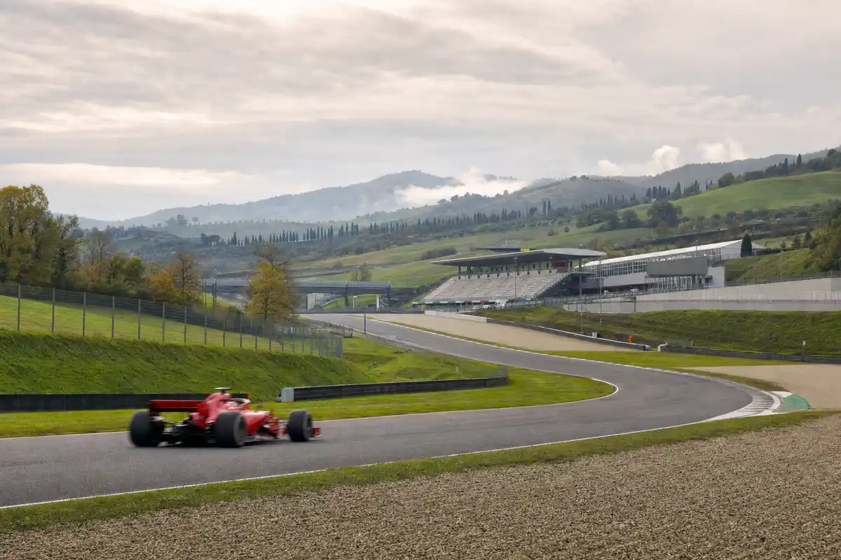 Monoposto rossa sfocata in accelerazione su una curva in salita, con tribune vuote e colline toscane sullo sfondo